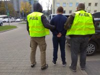 Policjanci ubrani w kamizelki odblaskowe stojący na parkingu policyjnym z zatrzymanym. Mężczyzna ma założone kajdanki na ręce trzymane z tyłu.
