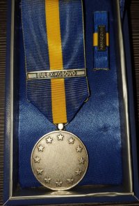 na stole , w ramce jest umieszczony złoty medal za zasługi w Kosowie , z niebiesko-żółtą przewieszką