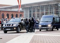 Zdjęcie jest zrobione przy rynku manufaktura, widać na nim dwa duże, ciemne samochody, obok nich biegną zamaskowani policjanci w kominiarkach i w mundurach.
