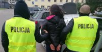 Zdjęcie jest zrobione na dziedzińcu Komendy Miejskiej Policji w Łodzi, w momencie kiedy kobieta wychodzi z policyjnego aresztu, w asyście dwóch policjantów.Zatrzymana kobieta, o szczupłej posturze, ciemnych długich włosach związanych w kucyk, ubrana w czarną kurtkę, jest prowadzona przez dwóch policjantów ubranych w żółte kamizelki policyjne z napisem POLICJA. Jeden z policjantów ma założony na głowę czarny kaptur . Kobieta ma założone kajdanki na ręce trzymane z tyłu.