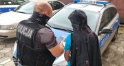 Nieumundurowany policjant w kamizelce z napisem POLICJA  stoi z zatrzymany sprawcą kradzieży przed radiowozem w dłoni trzyma odzyskany telefon komórkowy.