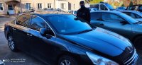 Na zdjęciu widoczny policjant w mundurze służbowym z maseczką na twarzy, który stoi przy odzyskanym pojeździe marki Peugeot. Pojazd koloru ciemny metalik.