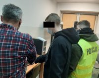 Zdjęcie jest zrobione również podczas badania odcisków palców, widzimy nam policjanta z zieloną kamizelką policyjną, zatrzymanego, oraz policjanta po cywilnemu, który pokazuje zatrzymanemu jak poprawnie ustawić dłonie.