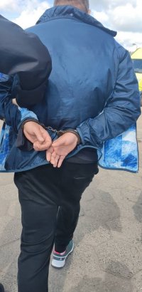 Policjanci prowadzą zatrzymanego mężczyznę po rynku. Mężczyzna ma założone kajdanki na ręce trzymane z tyłu.