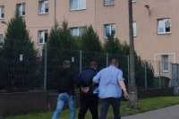 Policjanci prowadzą mężczyznę mającego założone kajdanki na ręce trzymane z tyłu do budynku Policji