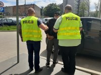 Mężczyzna jest prowadzony do cywilnego radiowozu policyjnego przez dwóch policjantów ubranych w żółte kamizelki policyjne. Ubrany jest w szare spodnie dresowe i ciemną kurtkę.