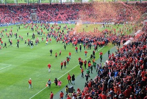 Wystrzelone konfetti na murawę boiska gdzie stoją piłkarze Widzewa Łódź. Na stadion wbiegają kibice.