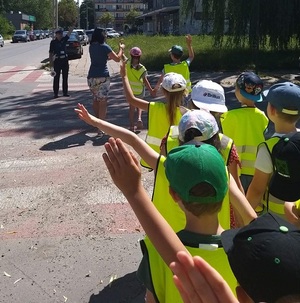Policjantka uczy dzieci prawidłowo przechodzić przez jezdnię, dzieci maja uniesione rączki.