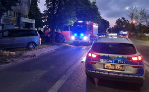 Miejsce wypadku, pojazdy policji i straży pożarnej z włączonymi światłami błyskowymi stoją na jezdni, w tle rozbity samochód.