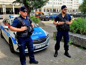 Na zdjęciu dwóch policjantów w umundurowaniu ćwiczebnym stojący na tle radiowozu i budynku w mieście. W oddali widoczna karetka pogotowia.
