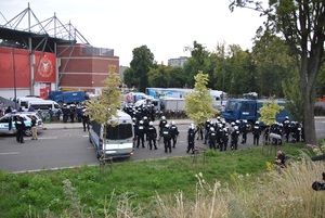 Policyjne zabezpieczenie przed stadionem. Policjanci stoją w grupach wokół stadionu.