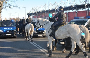 Służby podczas zabezpieczenia meczu, policjanci i strażnicy miejscy na koniach, w tle oznakowane radiowozy.