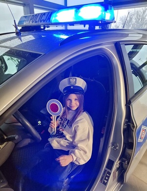 Dziewczynka siedzi w radiowozie w czapce policyjnej i z lizakiem w dłoni.