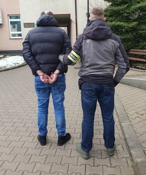 Nieumundurowany policjant prowadzi podejrzanego, który ma założone na rękach kajdanki.