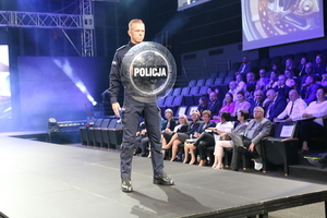 Policjant podczas pokazu mody służb mundurowych prezentuje umundurowanie , stoi na wybiegu, trzymając tarczę z napisem POLICJA, w tle widać publiczność.