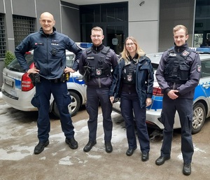 Policjant wraz ze studentami szkoły policyjnej z Niemiec stoją na tle radiowozów na dziedzińcu komisariatu.