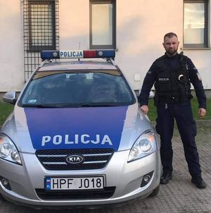 Umundurowany policjant stoi obok oznakowanego radiowozu.