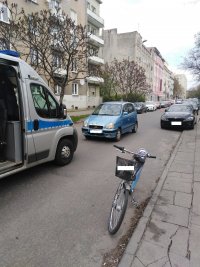 Na zdjęciu widoczny jest oznakowany radiowóz policyjny typu bus z otwartymi drzwiami bocznymi. Obok stoi rower z koszykiem. W tle pojazd koloru niebieskiego (fiat punto) oraz niskie zabudowania ciągnące się wzdłuż ulicy.