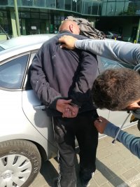 Na zdjęciu widoczny jest nieumundurowany policjant przytrzymujący zatrzymanego przy radiowozie. Mężczyzna trzymany przez policjanta ma założone kajdanki na ręce trzymane z tyłu.