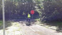 W tle widać rowerzystę ubranego w kamizelkę odblaskową koloru żółtego i kask rowerowy jadące po alejce pomiędzy drzewami i  zatrzymującego się przed znakiem STOP koloru czerwonego z białym napisem.