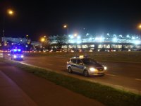 Na zdjęciu wykonanym w porze nocnej, widoczne są dwa oznakowane radiowozy policji, które jadą na pasie ruchu w okolicach stadionu miejskiego przy ul. Piłsudskiego w Łodzi. W tle widać rozświetlony stadion w trakcie trwania meczu.