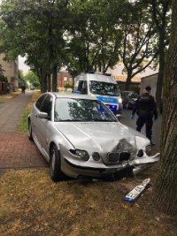 Samochód BMW jasnego koloru stoi przed drzewem z rozbitym przodem. W tle radiowóz policji i umundurowany policjant.