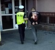 Policjant umundurowany z ruchu drogowego prowadzi zatrzymanego mężczyznę do budynku Komendy Miejskiej Policji w Łodzi.