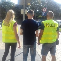 Na zdjęciu widzimy młodego szczupłego, mężczyznę, odwróconego tyłem do aparatu, ubrany jest w dżinsy i czarną koszulkę , po jego obu stronach widzimy policjantkę i policjanta , ubranych w żółte kamizelki z napisem policja.