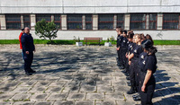 Z lewej strony zdjęcia widoczna postać policjanta w mundurze koloru granatowego. Z prawej strony zdjęcia widać dziesięć osób stojących obok siebie w jednym rzędzie. Osoby ubrane są w mundury koloru czarnego.
