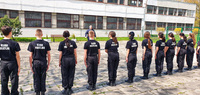 Z lewej strony zdjęcia widoczna postać policjanta w mundurze koloru granatowego. Z prawej strony zdjęcia widać dziesięć osób stojących obok siebie w jednym rzędzie. Osoby ubrane są w mundury koloru czarnego.
