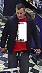 Mężczyzna w wieku około 30-40 lat, ubrany w ciemną kurtkę, czerwoną bluzę z białym napisem, szare spodnie dresowe i ciemne buty.
