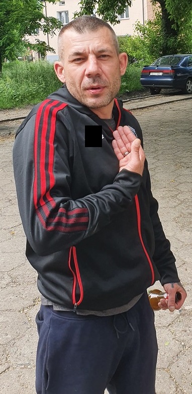 Mężczyzna w wieku około 25 lat, ubrany w koszulkę z krótkim rękawem koloru czerwonego, ciemne długie spodnie dresowe, ciemne buty sportowe oraz torbę koloru szarego.