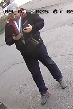  mężczyzna w wieku około 35-45 lat, szara czapka z daszkiem, granatowa kurtka z kapturem i czerwonym wnętrzem, szara koszulka, granatowe spodnie, biała smycz zawieszona na szyi, brak zarostu, krótkie włosy, na ramieniu zawieszona czarna saszetka.