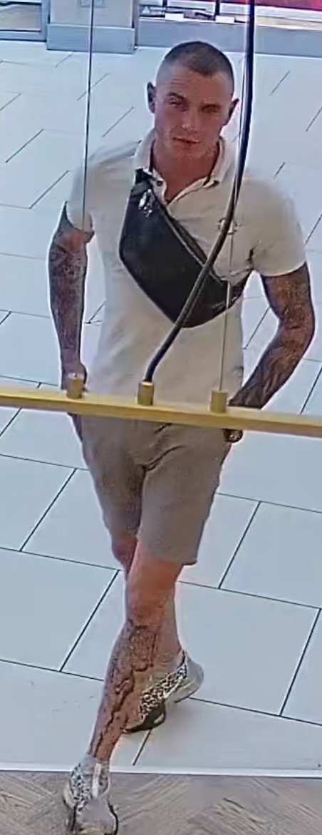  Mężczyzna w wieku około 25-35 lat, krótkie, czarne włosy, ubrany w białą podkoszulkę, szare spodenki o długości przed kolano, białe buty, przewieszona czarna saszetka, na obu przedramionach i nogach widoczne tatuaże.