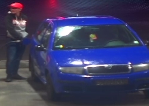 mężczyzna szczupłej budowy ciała, ubrany w czerwoną czapkę z daszkiem, czarno - szarą bluzę z białym poziomym paskiem, długie spodnie jeansowe w niebieskim odcieniu, czane buty z białą podeszwą.