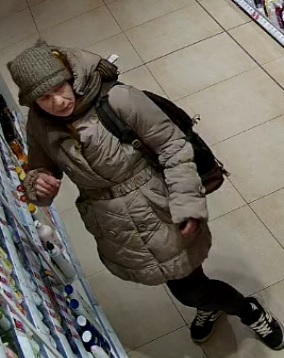 Kobieta w wieku około 40-50 lat, ubrana w beżową kurtkę z kapturem, czarne spodnie dresowe z elementami koloru białego, zieloną czapkę zimową, czarne buty sportowe z białymi elementami, przez ramię przewieszony brązowy plecak.