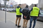 Na zdjęciu widzimy zatrzymanego mężczyznę , ubranego w ciemną kurtkę i beżowe spodnie. Mężczyzna stoi tyłem do aparatu , ma założone kajdanki z tyłu. Po jego obu stronach stoi dwóch policjantów w żółtych policyjnych kamizelkach.