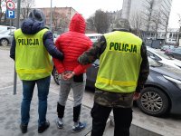 Na zdjęciu widzimy podejrzanego ubranego w czerwoną kurtkę , po obu stronach stoją policjanci w zielonych kamizelkach.