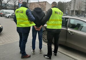 na zdjęciu widzimy zatrzymanego mężczyznę , którego trzymają po obu stronach policjanci ubrani w żółte kamizelki. Funkcjonariusze prowadzą zatrzymanego do radiowozu.