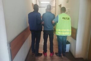 Na korytarzu stoi tyłem zatrzymany sprawca, którego po obu stronach trzymają policjanci.