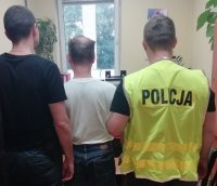 Na zdjęciu widzimy zatrzymanego mężczyznę, po jego obu stronach stoją policjanci, zdjęcie jest zrobione w pokoju przesłuchań.