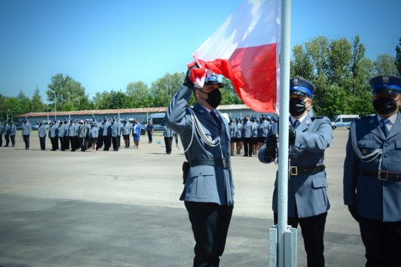 Policjanci w tle stojący w szyku, na pierwszym planie funkcjonariusze z flagą Polski stoją przy maszcie