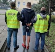 Zatrzymany stoi w krótkich spodenkach przy policyjnej izbie zatrzymań na dziedzińcu Komendy Miejskiej Policji w Łodzi, ma założone kajdanki z tyłu, po jego obu stronach stoi dwóch policjantów ubranych w żółte kamizelki.