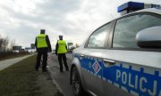 Policjanci ruchu drogowego stoją przy jezdni, kontrolują zachowanie uczestników ruchu drogowego,  w tle stoi zaparkowany oznakowany radiowóz