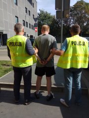 Policjanci w kamizelkach z napisem POLICJA eskortują zatrzymanego sprawcę pobicia.