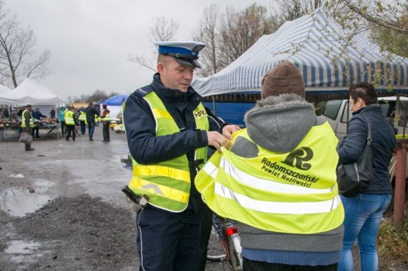 Umundurowany policjant ruchu drogowego na targowisku pomaga założyć kamizelkę odblaskową rowerzystce. W tle widać inne osoby ubrane w odblaskowe kamizelki i stragany.