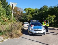 Umundurowani policjanci ruchu drogowego stoją na drodze  przy oznakowanym radiowozie, za nimi stoi srebrny samochód, którego kierujący przekroczył prędkość.