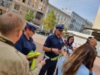 Na zdjęciu widoczna jest dwójka policjantów ruchu drogowego. Policjanci ubrani są w niebieskie mundury i białe czapki. Funkcjonariusze udzielają wywiadu grupie dziennikarzy na ul. Piotrkowskiej w Łodzi. Policjanci trzymają w rękach elementy odblaskowe, które mają być rozdawane przechodniom. W tle widać kamienice i fragment deptaka ulicy Piotrkowskiej.