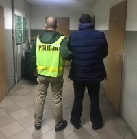 Zdjęcie jest zrobione na korytarzu V Komisariatu Policji, policjant, ubrany w cywilne ubranie ma założoną kamizelkę policyjną , z napisem Policja. Trzyma za rękę zatrzymanego , ubranego w granatową kurtkę i ciemne spodnie . Ma ciemne włosy