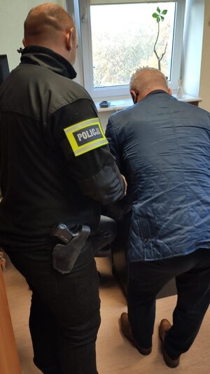 Policjant w ubiorze cywilnym z przepaską na ramieniu z napisem "policja" stojący w pomieszczeniu za zatrzymanym mężczyzną. Zatrzymany ubrany w niebieską pikowaną kurtkę i czarne spodnie.